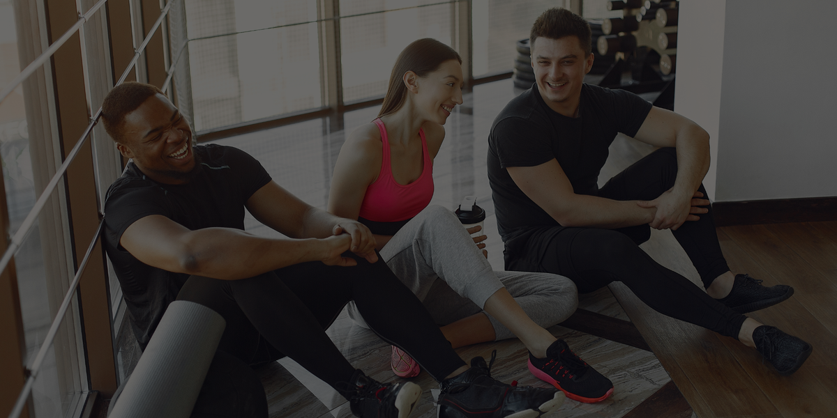 Three people sitting together in a gym or fitness space, wearing athletic clothing. The scene is somewhat dim and shadowed, with the central figure wearing a pink sports top and light gray leggings.